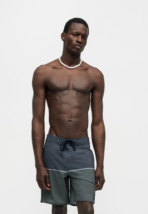 Uomo senza maglietta che indossa una collana bianca di perle e pantaloncini da bagno grigio scuro con coulisse, in piedi davanti a uno sfondo chiaro e uniforme.