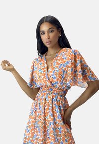 Bloemendress met oranje en blauwe patronen, korte vlindermouwen, een diepe V-halslijn en een gesmokte taille voor een A-lijn silhouet.