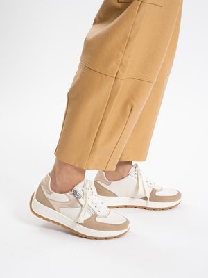 Person wearing beige cropped pants and white sneakers with beige and tan accents, standing on a white background.