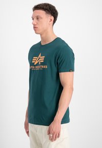 Teal bomulds T-shirt med korte ærmer, der har en orange grafisk "ALPHA INDUSTRIES" logo og et lille sort mærke på ærmet.