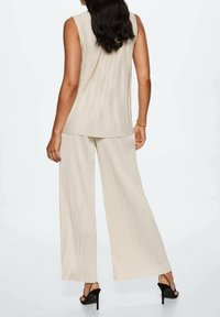 Beige plisovaný top a kalhoty se širokými nohavicemi, vyrobené z lehkého materiálu. Oblečení má béžovou barvu a hladkou texturu. Černé vysoké podpatky.