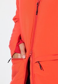 Czerwona kurtka z zamkiem błyskawicznym o gładkiej fakturze, wyposażona w kieszenie i regulowane suwaki. Wnętrze w kontrastowym kolorze.