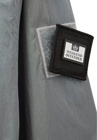 Tessuto di giacca grigia con una texture liscia, caratterizzato da una toppa tessuta nera con l'etichetta "WEEKEND OFFENDER," bordata in finta pelliccia grigia.