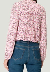 Kobieta ubrana w bluzkę z długim rękawem w białym kolorze z różowym kwiecistym wzorem i plisowanym tyłem, w połączeniu z dżinsami z wysokim stanem w niebieskim kolorze.