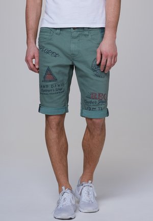 Man draagt teal korte broek met grafisch patroon en opgerolde pijpen, witte sneakers en een wit shirt, staand tegen een effen grijze achtergrond.