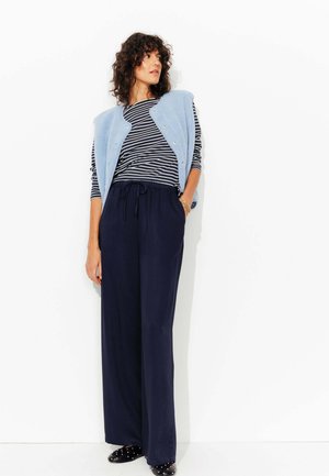 Un gilet sans manches bleu clair sur une chemise à manches longues noire et blanche à rayures, associé à un pantalon large marine et des chaussures noires à enfiler.