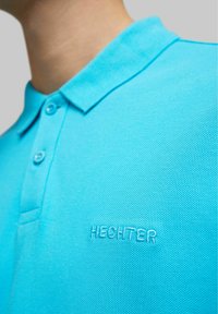 Turquoise poloshirt van gestructureerde stof met een kraag, voorzien van twee knopen en een geborduurd logo met de tekst "HECHTER" op de voorkant.