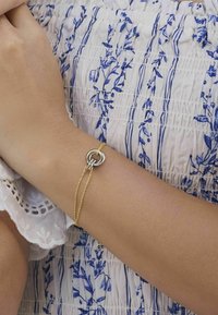 Armband av guld och silver med två sammanflätade ringar, bärs på ett handled. I bakgrunden syns en ljus blommönstrad tyg.