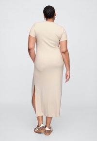 Beżowa, do kolan, dopasowana sukienka wykonana z miękkiego materiału, z krótkimi rękawami i bocznym rozcięciem, zestawiona z casualowymi brązowymi butami.