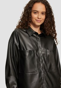 Zwarte faux leren shirt met knoopsluiting aan de voorkant, grote borstzakken en een klassieke kraag. Gladde textuur met een relaxte pasvorm.