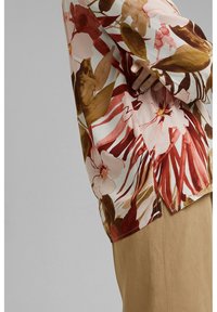 Blusa floreale in tessuto leggero con grandi fiori rosa e marroni e foglie verdi, con spacchi laterali e vestibilità ampia, abbinata a pantaloni beige.