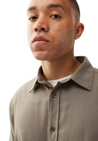 Beige skjorte med krave, tekstureret stof og to synlige knapper. En hvid t-shirt er lagd under.