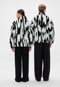 Műszőrme kabát fekete-fehér absztrakt mintával, magas gallérral és laza fazonnal, széles fekete nadrággal párosítva.