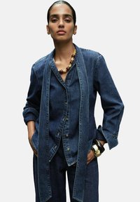 Denim jacket with long, draped front, button closure, and rolled sleeves. Paired with a textured bracelet and a beaded necklace.