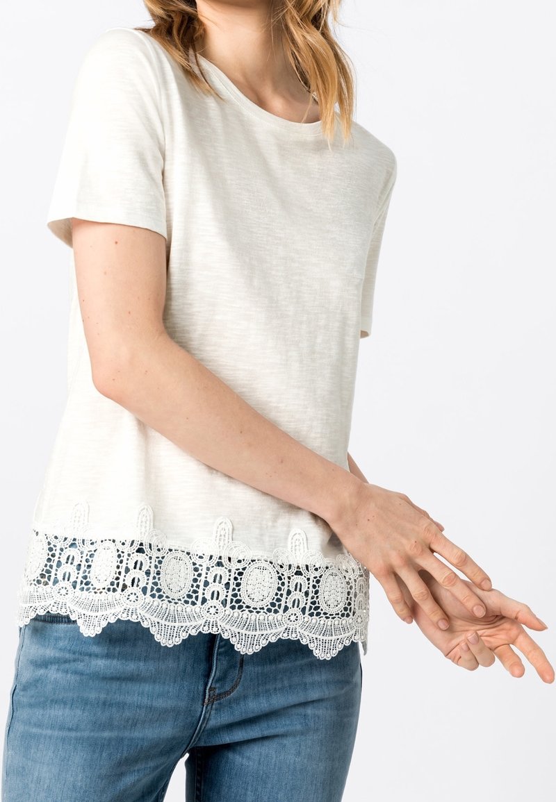 Top corto de manga corta en beige claro con un dobladillo de encaje texturizado y patrones intrincados. Combinado con unos jeans de denim azules, mostrando un estilo casual.