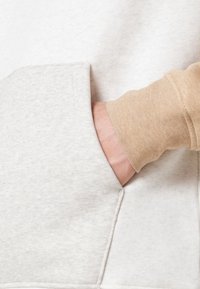 Main dans une manche beige insérée dans la poche avant d'un sweat-shirt gris clair, montrant la texture du tissu et les détails des coutures de la poche.