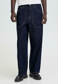 Donkerblauwe denimbroek met rechte pijpen, contrasterende witte stiksels, meerdere zakken en een comfortabele pasvorm. Taille voorzien van een knoopsluiting.