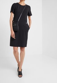 Robe noire à manches courtes avec une taille cintrée et des poches, associée à des chaussures noires à bout ouvert et un petit sac noir. Texture lisse.