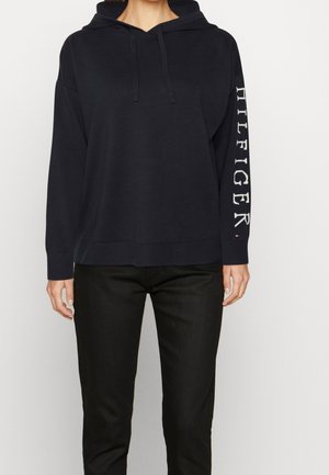 Personne portant un sweat à capuche noir avec le texte "HILFIGER" sur la manche droite et un pantalon noir, visible du cou aux hanches sur un fond blanc uni.
