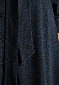 Kratké rukávy šaty v tmavomodrom materiáli s textúrovaným leopardím vzorom. Obsahuje gombíky a viazací akcent na páse.