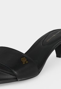 Μαύρο πέδιλο με ανοιχτά δάχτυλα και χαμηλό τακούνι τύπου block, χρυσό λογότυπο "TH" στο λουρί και ένδειξη Tommy Hilfiger στην εσωτερική σόλα.