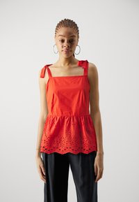 Rød bomulls peplum-top med firkantet utringning, knyteseler og utskåret broderi i kanten. Brukt med mørke bukser.