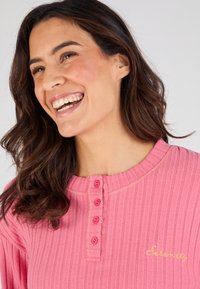 Top de manga larga de canalé rosa con un escote tipo henley y cinco botones rosa, con texto bordado en dorado que dice "Serenity" cerca del dobladillo.