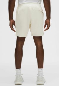 Hombre vistiendo pantalones cortos deportivos color blanco hueso, calcetas blancas y zapatillas blancas, de pie con las piernas y brazos visibles desde atrás sobre un fondo liso.