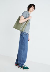 Olivengrüner Baumwoll-Tote-Bag mit großem "ATOMIC"-Logo. Kombiniert mit einem grauen T-Shirt und lockeren blauen Denim-Jeans, die grüne Turnschuhe tragen.