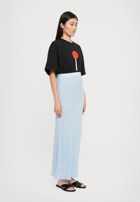 Kvinna i en svart oversized t-shirt med röd lollipopgrafik, ljusblå långkjol och svarta slip-on sandaler, som står mot en vit bakgrund.