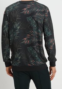 Uomo che indossa una maglietta a maniche lunghe nera con motivi di foglie tropicali verde scuro e rosa, visto da dietro, in piedi davanti a uno sfondo semplice.