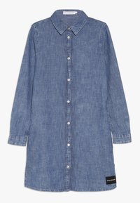 Džínová košilová šaty v středně modr é barvě s knoflíkovým zapínáním, špičatým límcem, dlouhými rukávy a černou štítkovou akcentací na spodním lemu.