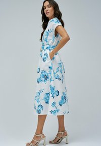 Robe maxi florale en coton blanc avec des motifs de fleurs bleues, poches latérales, manches courtes et taille cintrée, assortie à des sandales à lanières.
