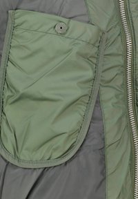 Kurtka ocieplana z zielonym zewnętrznym materiałem i dużą kieszenią na guziki. Tkanina jest gładka z subtelną fakturą, uzupełniona srebrnym zamkiem błyskawicznym.