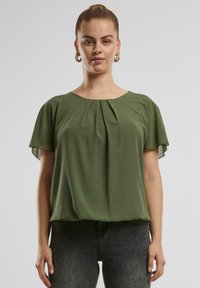 Grüne kurzärmelige Bluse aus leichtem Stoff, mit Rundhalsausschnitt und plissierter Vorderseite. Kombiniert mit dunkelgrauen Jeans.