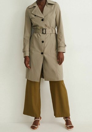 Kvinde iført en beige trenchcoat med bælte og sorte knapper, olivengrønne vidde ben-bukser og hvide sandaletter med hæl mod en ensfarvet væg.