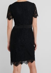 Schwarzes Spitzenkleid mit kurzen Ärmeln, gewelltem Saum und taillierter Taille, die durch ein breites schwarzes Band betont wird. Der Reißverschluss auf der Rückseite ist sichtbar.