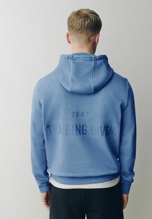 Mann mit dem Rücken zur Kamera trägt einen blauen Kapuzenpullover mit der Aufschrift "7841 TRAINING DIVS.N." auf dem Rücken vor einfarbigem Hintergrund.