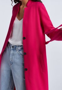 Fuksia-villasekoite pitkä takki suoralla leikkauksella, nappikiinnityksellä ja sivutaskulla; käytetty valkoisen topin ja vaaleansinisten farkkujen päällä.