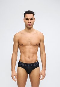 Μυώδης άνδρας όρθιος, φορώντας μαύρα εσώρουχα Calvin Klein, μπροστά σε απλό ανοιχτό γκρι φόντο, κοιτάζοντας προς τα εμπρός με ουδέτερη έκφραση.