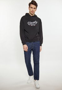 Černá mikina s kapucí s bílým potiskem "Varsity" a "since 1998". Vybaveno modrými džíny a bílými teniskami. Jednoduché pozadí.