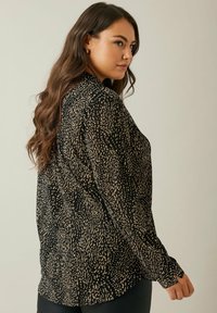 Schwarze Bluse mit einem strukturierten beige Muster, mit langen Ärmeln und einem abgerundeten Saum. Das Material wirkt leicht und halbtransparent.