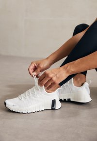 Weiße Sportschuhe mit strukturiertem Obermaterial, flexibler Sohle und schwarzen Akzenten. Die Schnürsenkel werden gebunden, wodurch der Sitz und das Design des Schuhs hervorgehoben werden.