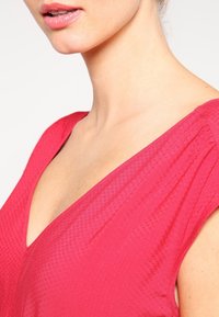 Haut fuchsia avec un décolleté en V profond, tissu texturé et détails plissés aux épaules. Fini lisse et couleur vibrante améliorent l'attrait visuel.