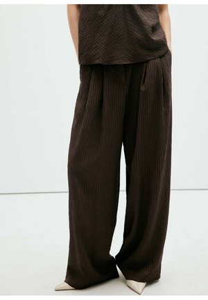 Persona que lleva pantalones anchos y sueltos de textura marrón oscuro y parte superior a juego, con zapatos blancos puntiagudos, de pie sobre un fondo claro y liso.
