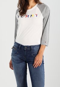 Weißes Hemd mit grauen Raglanärmeln, auf dem der Text "TOMMY" in bunten Buchstaben zu sehen ist; kombiniert mit dunkelblauen Jeans.