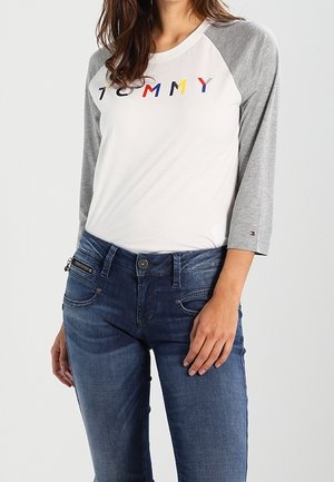 Camicia bianca con maniche raglan grigie che presenta la scritta "TOMMY" in lettere multicolori; abbinata a jeans in denim blu scuro.
