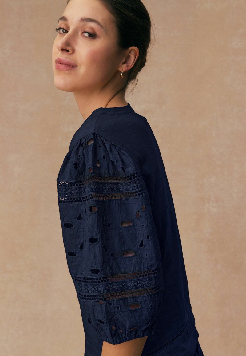 Jonge vrouw met donker gevlochten haar draagt een marineblauwe blouse met kant en halsuitsnijding en pofmouwen, kijkt over haar schouder tegen een beige achtergrond.