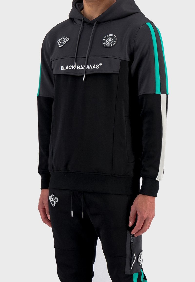 Conjunto de sudadera con capucha negra y pantalones de chándal que presenta un diseño de bloques de color gris y negro, acentos en turquesa, logos bordados y detalles de bolsillos con cremallera.