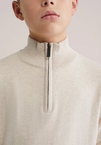 Šviesiai smėlio spalvos megztinis su užtrauktuku, aukštu apykakliu, tvirtu raštu ir juodu užtrauktuku. Audinys atrodo minkštas ir lengvas.